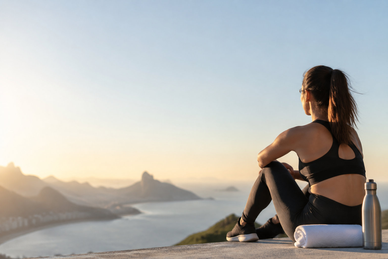 Une femme en tenue de sport observe un paysage côtier au coucher du soleil, assise après un entraînement.